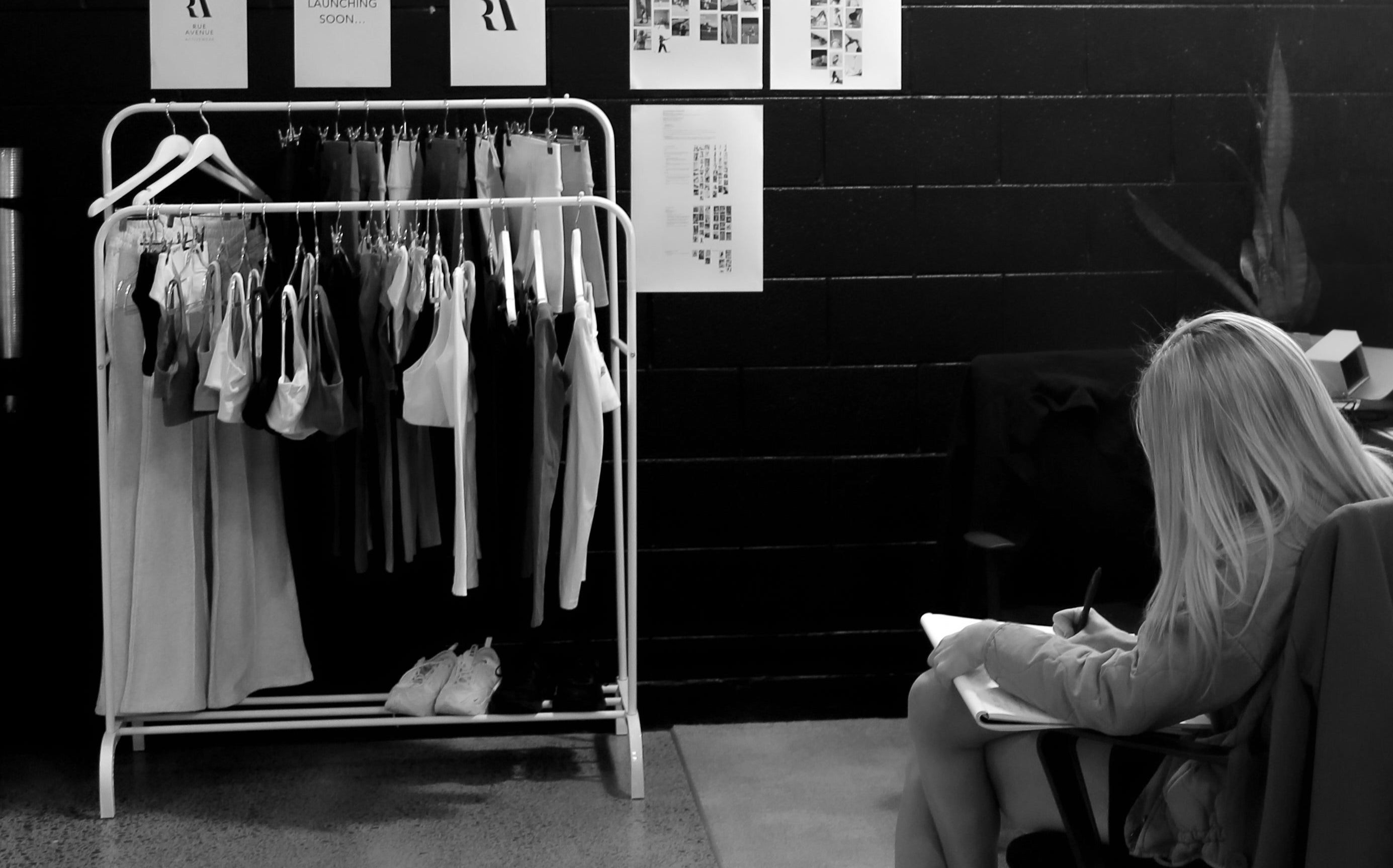 Rue Avenue founder sitting and writing next to a rack of clothes against a black wall.