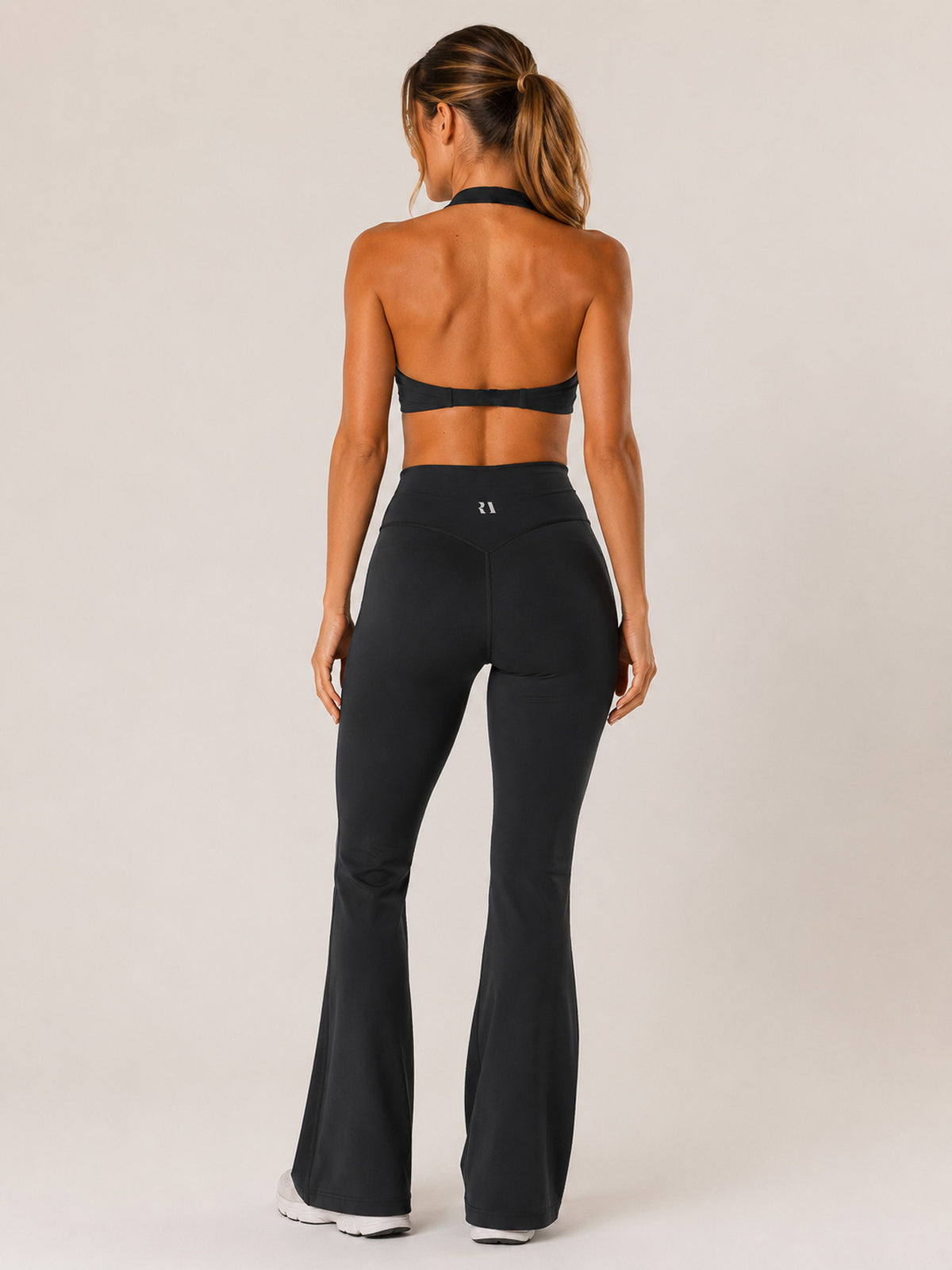 A woman shown from the back wearing black high-waisted flared leggings and a matching black halter-style sports bra against a plain light beige studio background.