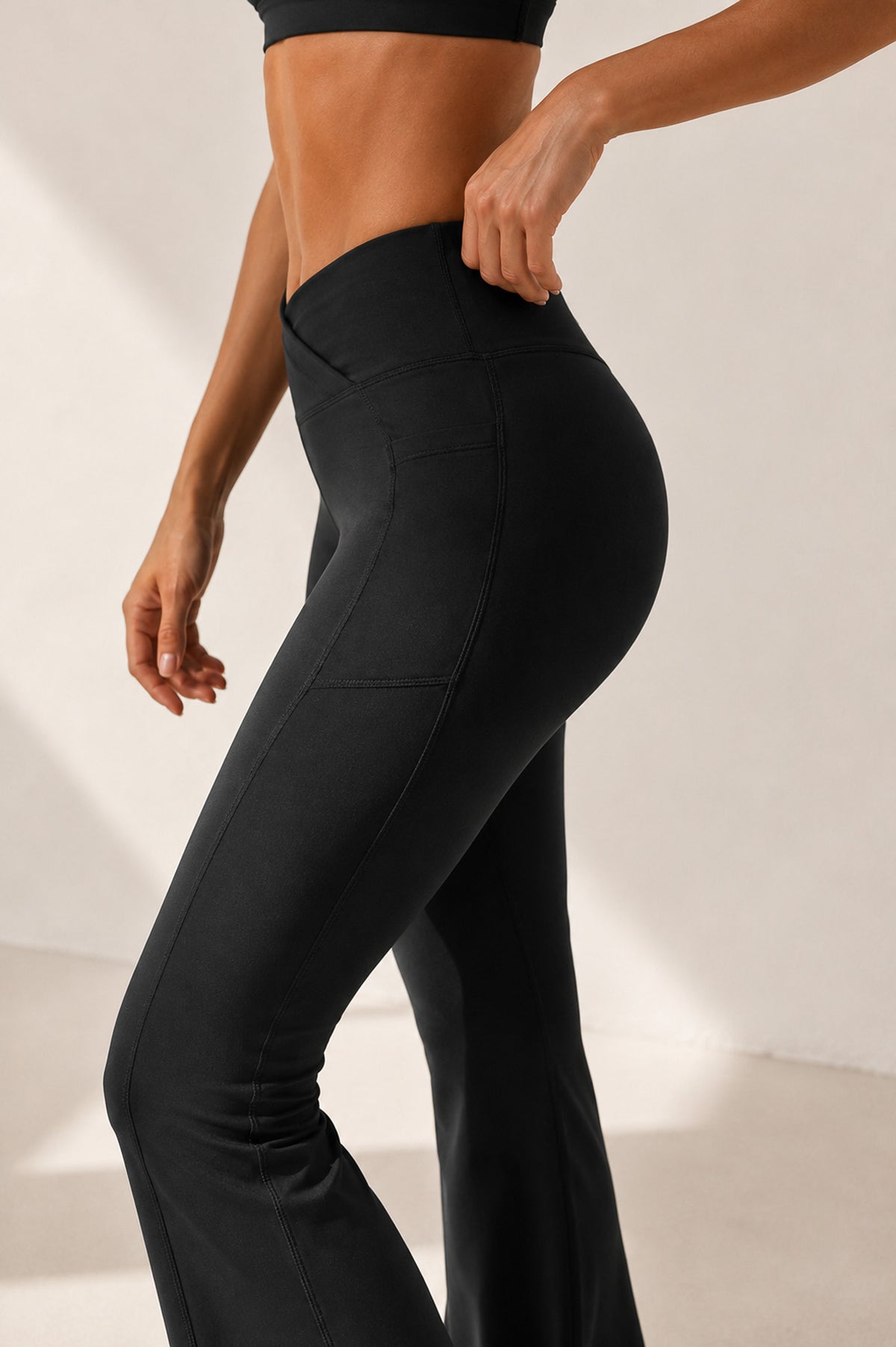 A close-up side view of a model wearing black high-waisted flared leggings, showing the side pocket and fit against a warm beige studio background.