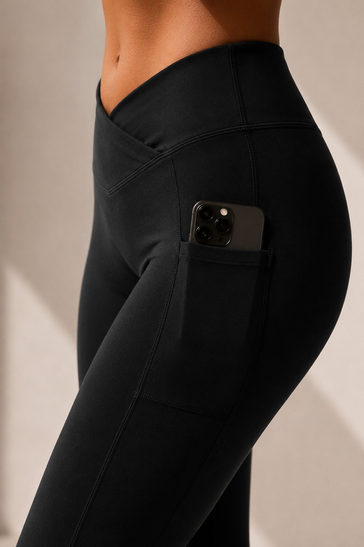 Close-up of a person wearing black high-waisted leggings with a side pocket holding a smartphone, photographed against a warm beige wall.